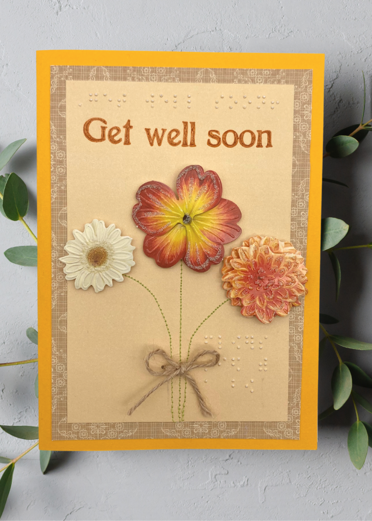 a vibrant orange card with 3 flowers tied with a twine ribbon that says get well soon. The card sits on a neutral grey backdrop