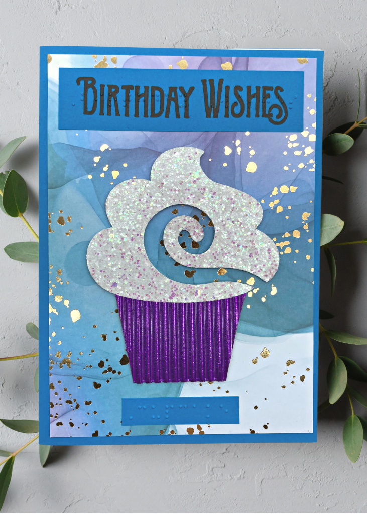 a bright blue card that says birthday wishes with a sparkly cupcake in a shiny corrugated cupcake tin on a multi-coloured blue, purple and teal background with gold flecks. The card sits on a neutral grey backdrop