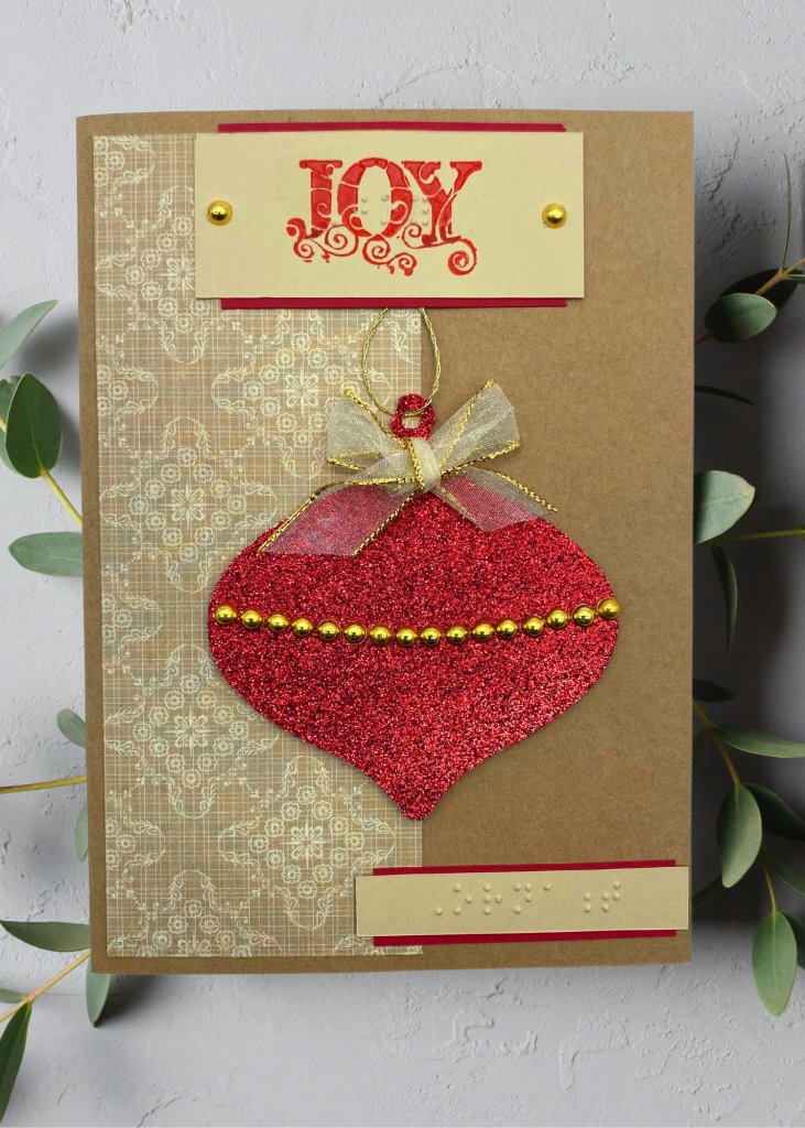 a brown card with bright red hanging ornament which says Joy. The card sits on a neutral grey backdrop