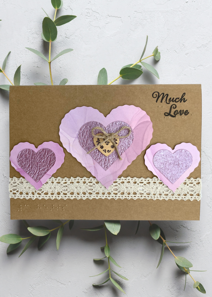a neutral brown card with nesting pink hearts that says Much Love on a neutral grey background