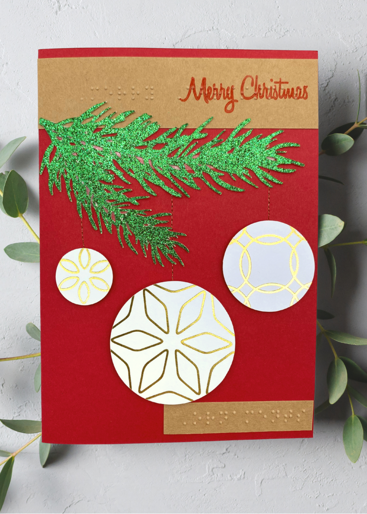 A red card with brown accents that says Merry Christmas with a green evergreen bough and 3 hanging ornaments on a neutral grey background.