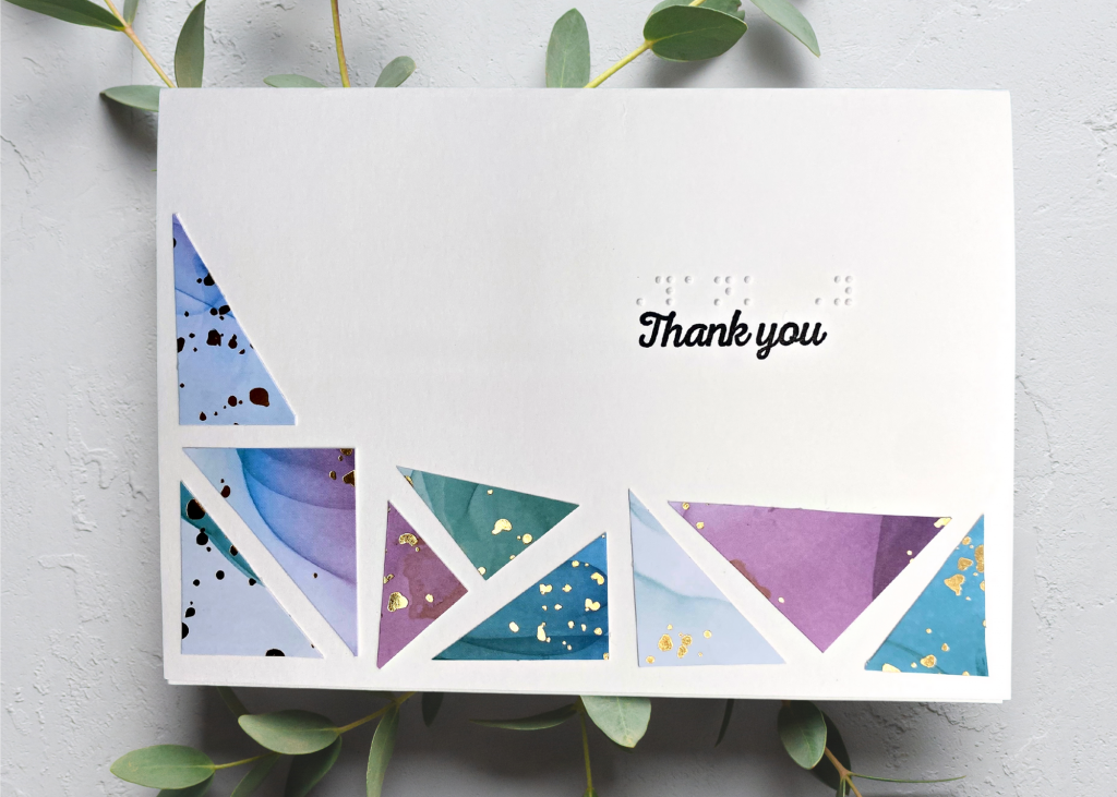 A white thank you card with many multi-coloured triangles creating a mosaic border. The card is sitting on a natural grey backdrop