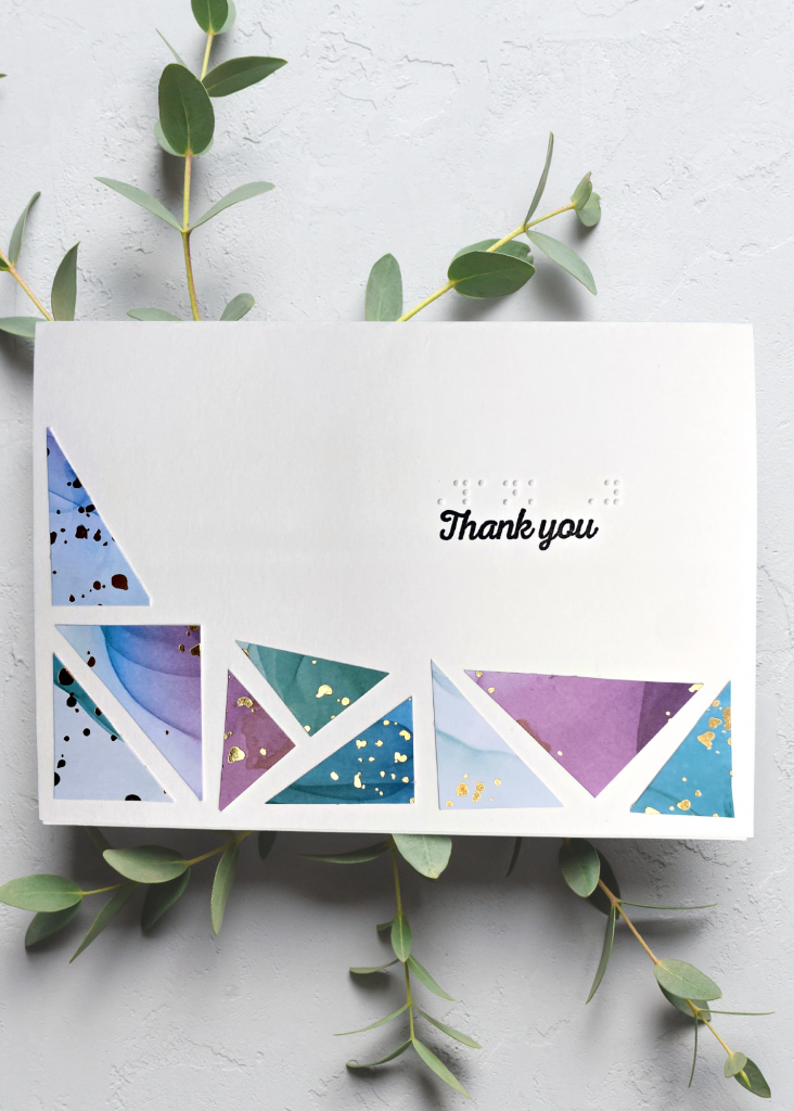 a white thank you card with a border of different sized multi-coloured triangles creating a mosaic on a neutral grey background