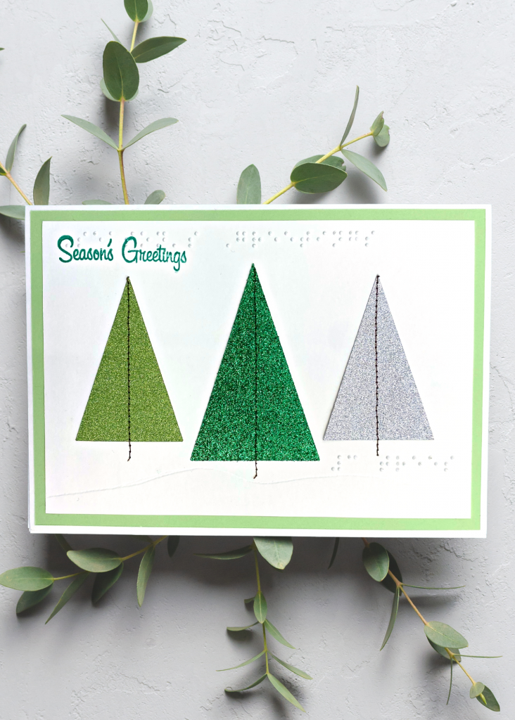a white and green card with 3 triangular trees in the snow that says Season's greetings. The card sits on a neutral grey backdrop