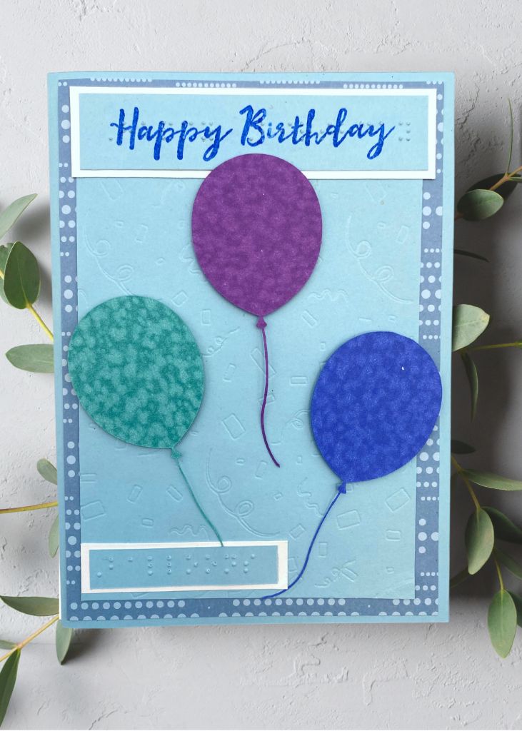 A blue card that says happy Birthday with 3 balloons on a neutral grey background.