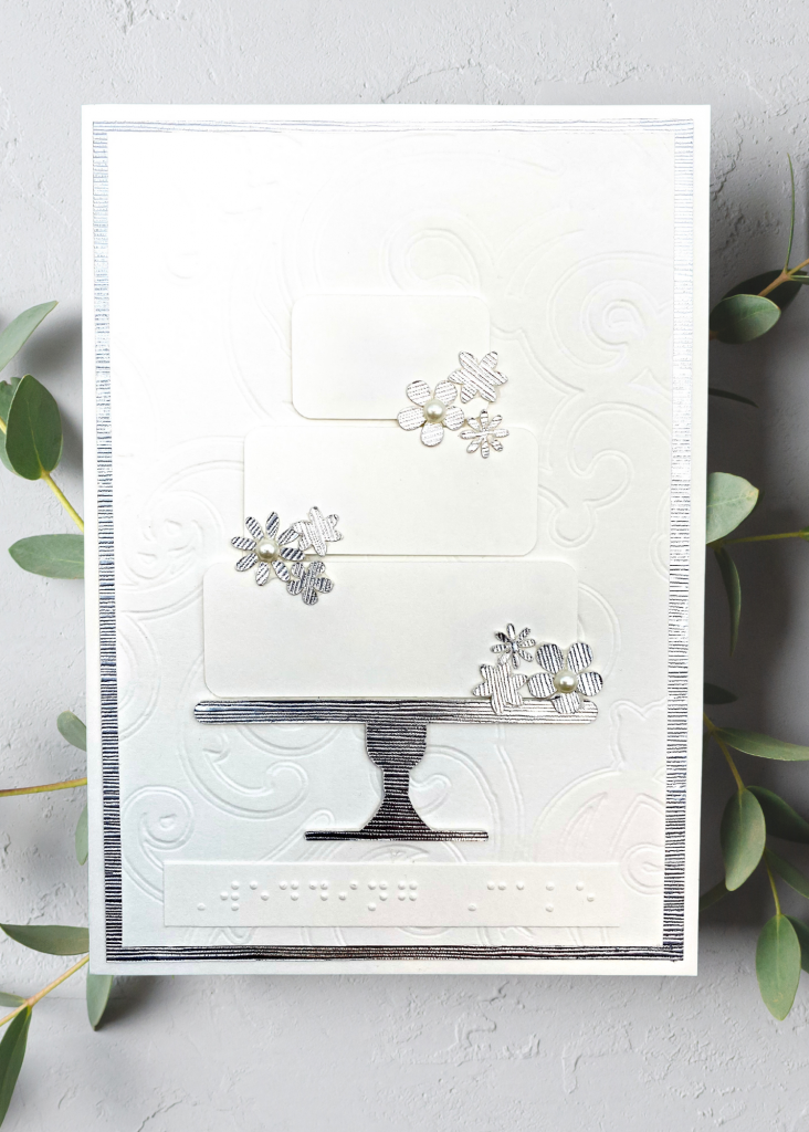 a white and silver card with a 3-tiered white wedding cake on a silver stand decorated with silver flowers and pearls. It sits on a neutral grey backdrop