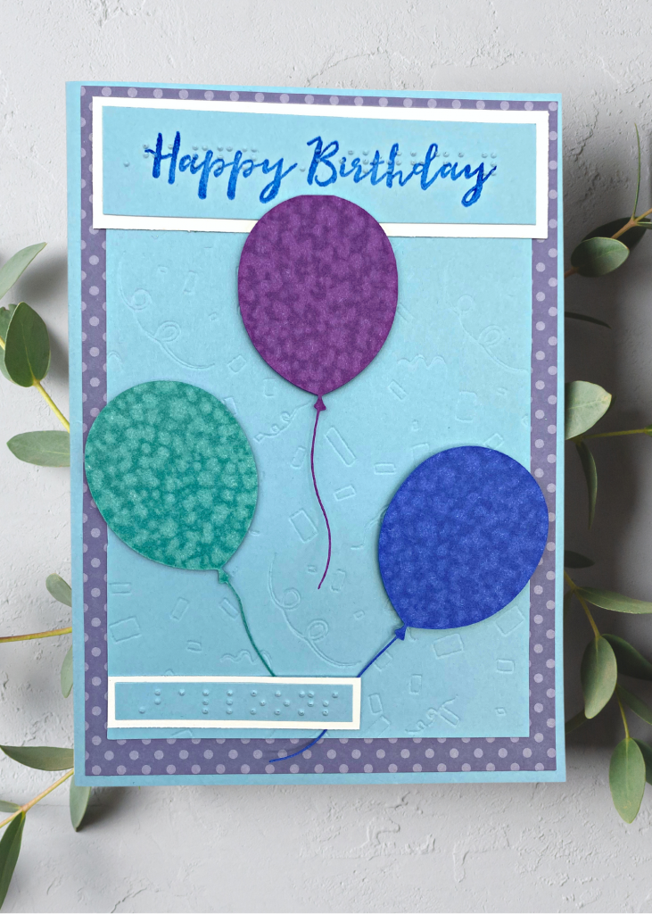 a light blue and purple polkadot card with 3 balloons floating on a textured confetti background. The card says Happy Birthday. The card is sitting on a neutral blue background