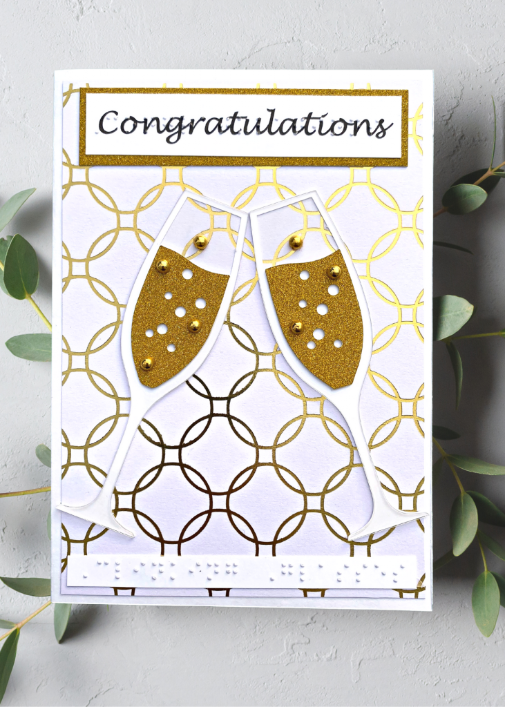 a white and gold card with bubbly champagne glasses clinking. Text says Congratulations. The card is sitting on a neutral grey backdrop