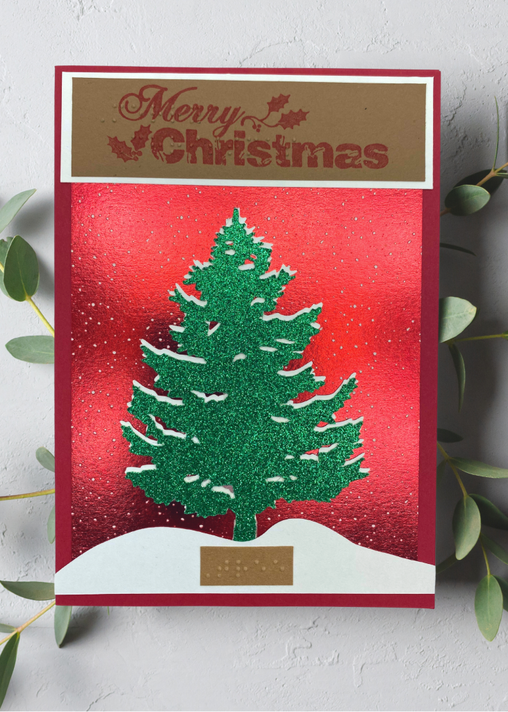a red card with a snowcapped evergreen tree sitting in the snow. The card says Merry Christmas. The card is sitting on a neutral grey backdrop