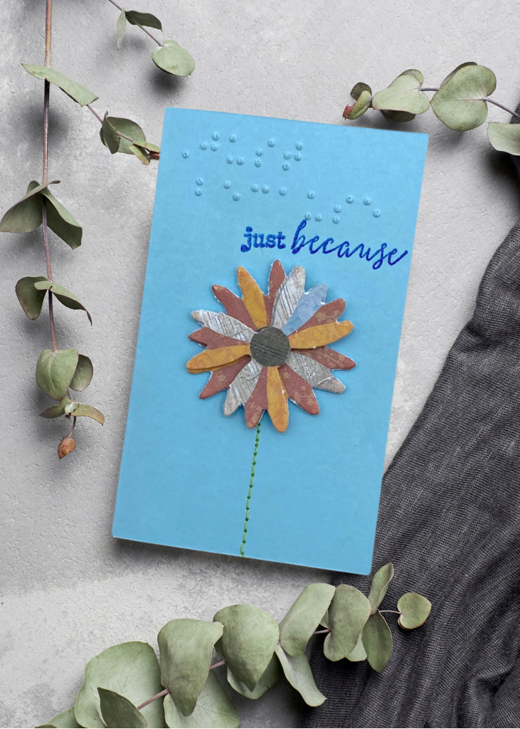 A pale blue card with a multi-coloured daisy that says just because sitting on a neutral grey backdrop