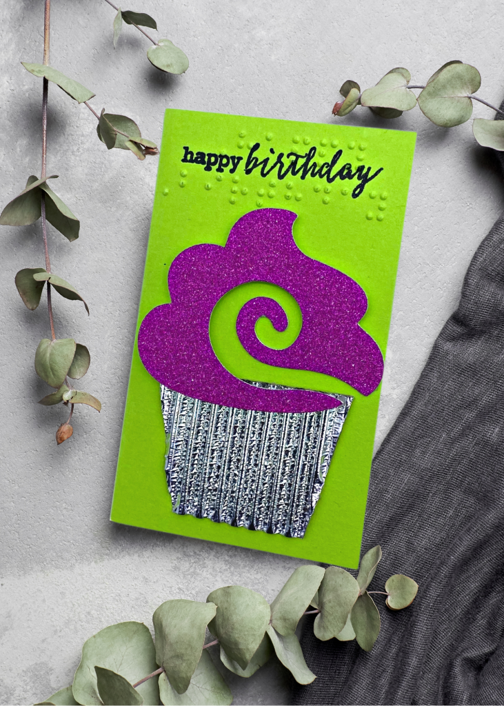 a neon green card that says happy birthday with a fuchsia cupcake in a silver tin on a neutral grey background