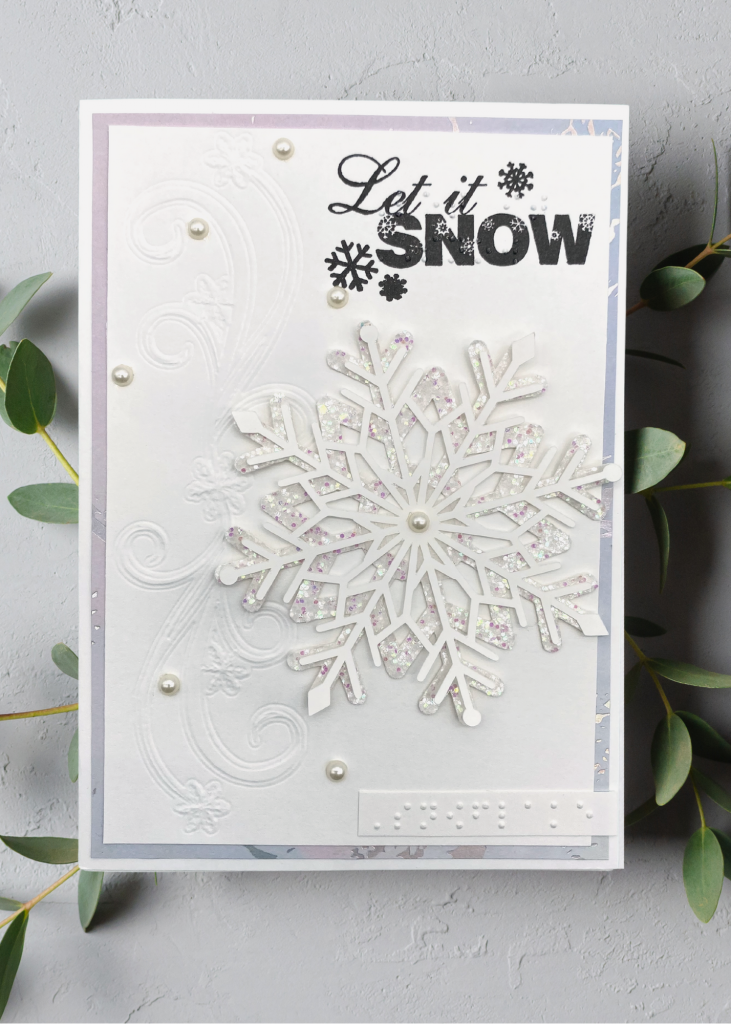 a white card with embossed and raised snowflakes that says Let it snow. The card sits on a neutral grey backdrop