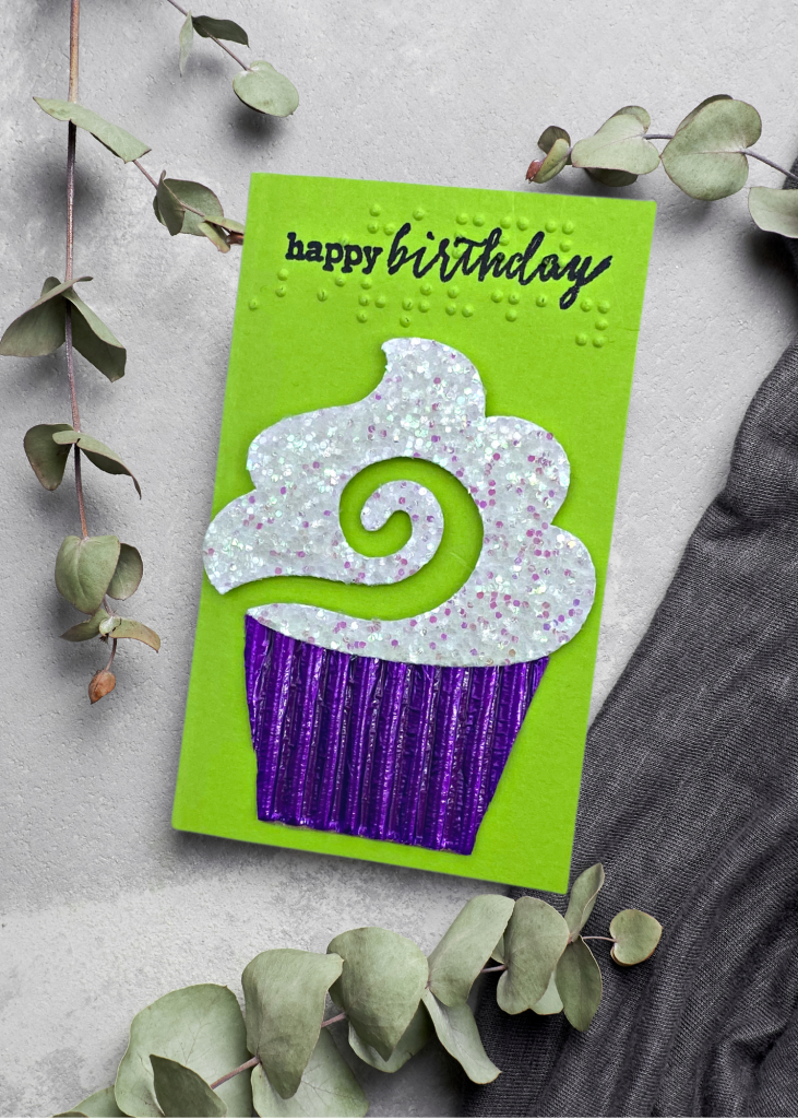 a neon green card with text that says Happy birthday and a sparkly iced cupcake in a purple tin on a neutral grey background